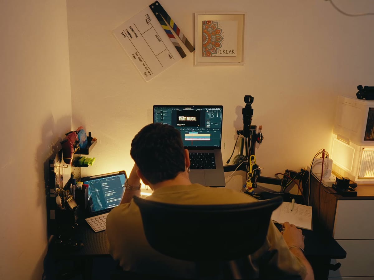 A man seen from behind, working at two screens at a desk in a warmly lit room
